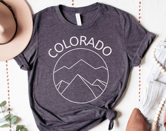 Op de afbeelding: Een donkergrijs T-shirt met een witte afbeelding van bergen en het woord "COLORADO" in een cirkel. Het shirt ligt op een wit oppervlak met een hoed, jeans en schoenen.