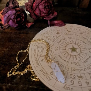 May include: A wooden circular divination board with a gold chain and a white crystal pendant. The board has a star in the centre and the days of the week and months of the year around the edge. The board is surrounded by dried roses.