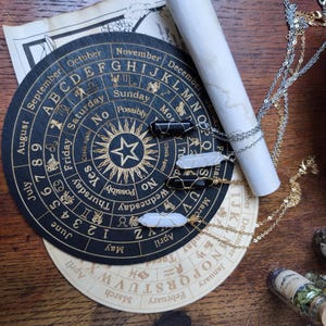 Pode incluir: Um tabuleiro de adivinhação preto e dourado com um design de sol e estrela. O tabuleiro tem os dias da semana, os meses do ano e os números de 1 a 9. Três colares com pingentes de cristal branco e preto estão em cima do tabuleiro.