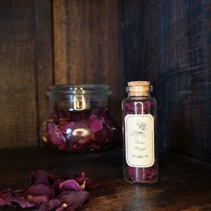May include: A glass jar and a small glass bottle filled with dried rose petals. The bottle has a cork stopper and a label that reads "Rose Rouge".