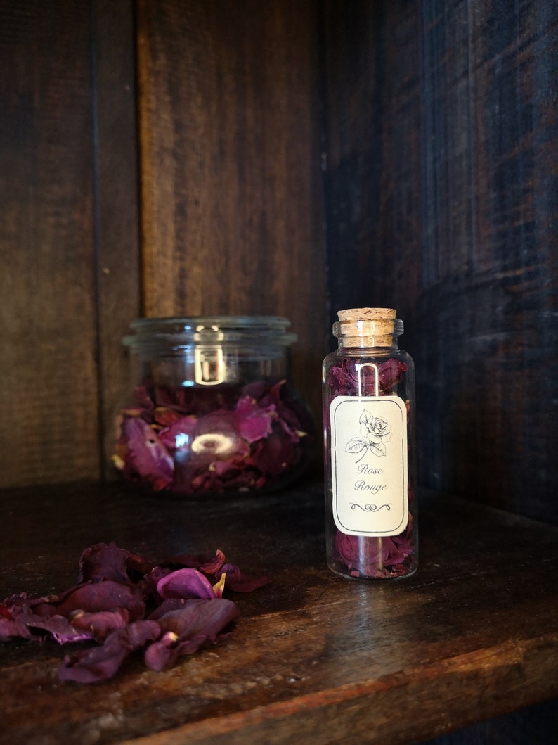 May include: A glass jar and a small glass bottle filled with dried rose petals. The bottle has a cork stopper and a label that reads "Rose Rouge".