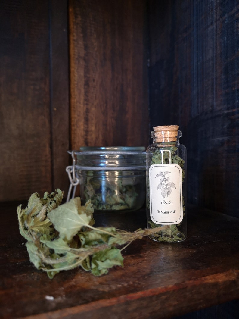 May include: A glass jar and a small glass bottle with a cork stopper, both containing dried herbs. The bottle has a label that reads "Ortie". The jar and bottle are sitting on a wooden shelf.
