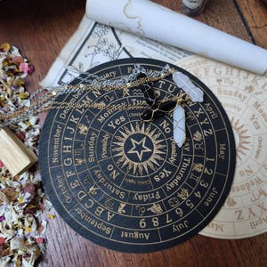 Op de afbeelding: Een zwart en gouden cirkelvormig divinatiebord met een ster in het midden en dierenriemsymbolen rond de rand. Het bord is omringd door kristallen en een opgerolde kaart.