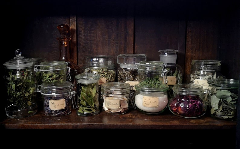 May include: A wooden shelf with a variety of glass jars filled with dried herbs and flowers. The jars have labels with handwritten text.