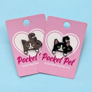 May include: Two enamel pins on pink cards with white hearts. The pins are of black cats with white markings, one with brown fur and the other with black fur. The text on the cards reads "Pocket Pet Collectible Enamel Pin".