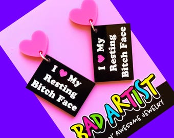May include: A pair of black acrylic earrings with pink hearts and the text "I  My Resting Bitch Face" on each earring. The earrings are displayed on a pink background with the text "BAD ARTIST FREAKIN' AWESOME JEWELRY" in rainbow colors.