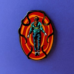 May include: A black and red enamel pin featuring a person standing in a swirling, red and orange portal. The person is wearing a blue shirt, black pants, and has brown hair.