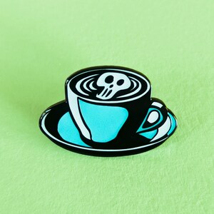 May include: A black and white enamel pin of a coffee cup with a skull in the foam. The cup is on a black saucer.