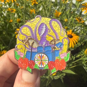 Puede incluir: Un colorido pin de esmalte con un diseño estilizado con un pulpo morado, una cámara azul y rosas rojas. El pin tiene un fondo amarillo de vidrieras y un diseño de rosa de los vientos. El pin se sostiene frente a flores amarillas.