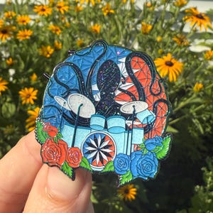 Puede incluir: Un colorido pin de esmalte con un pulpo tocando la batería. El pin tiene un esquema de color azul, rojo y blanco con un borde floral de rosas rojas y azules. La batería es azul claro con un diseño de estrella blanca.