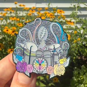 Puede incluir: Un pin de esmalte colorido que representa un pulpo tocando una batería. El pulpo es gris con tentáculos rosas, azules y grises. La batería es gris con un centro de colores del arcoíris. El pin está rodeado de rosas coloridas.