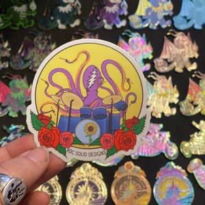 May include: A sticker featuring a purple octopus playing drums with a sunflower in the center. The sticker has a yellow background with red roses around the octopus. The text "ROC SOLID DESIGNS" is at the bottom of the sticker.