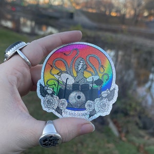 May include: A silver glitter sticker with a rainbow holographic background featuring a cartoon octopus playing drums. The octopus has a lightning bolt on its head. The sticker has a white outline and the text "OG SOLID DESIGNS" at the bottom.