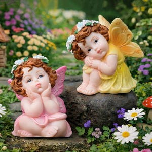 May include: Two fairy figurines, one in pink and the other in yellow, are seated outdoors. The fairies have brown hair, flower crowns, and wings. The pink fairy is in a pink dress, and the yellow fairy is in a yellow dress. The background features flowers and greenery.
