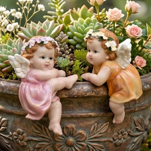 Op de afbeelding: Twee cherubijnachtige beeldjes met vleugels en bloemenkransen sieren een decoratieve plantenbak gevuld met vetplanten en rozen. Eén cherubijn draagt een roze jurk, de andere een oranje jurk. De plantenbak heeft een bronzen afwerking met bloemendetails.