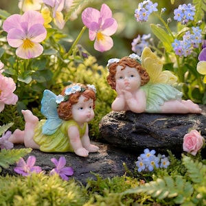 Peut inclure: Deux figurines de fées aux cheveux roux, couronnes de fleurs et ailes jaunes et vertes, posées sur un rocher sombre. Les fées sont entourées de fleurs colorées, dont des pensées roses et violettes, et des myosotis bleus, créant une scène de jardin fantaisiste.