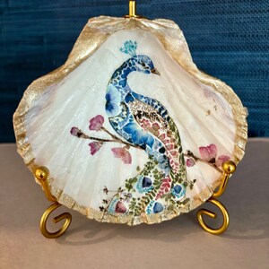 May include: A decorative seashell featuring a painted peacock design. The peacock is rendered in shades of blue, green, and pink, perched on a branch with pink blossoms. The shell has a gold-painted edge and is displayed on a gold metal stand.