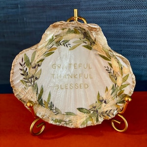 May include: A decorative seashell with gold trim and a gold stand. The shell features the words "Grateful, Thankful, Blessed" in gold lettering, surrounded by a wreath of green leaves and gold accents.