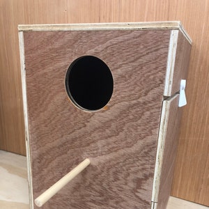 May include: A wooden birdhouse with a round entrance hole and a removable front panel. The birdhouse has a small wooden perch attached to the front.