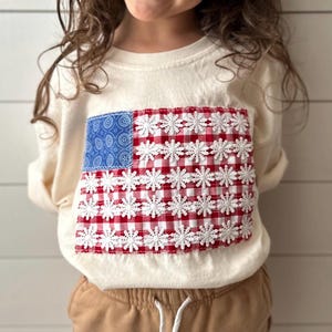 May include: Cream-colored long-sleeved shirt featuring a patriotic American flag design. The flag is made of blue patterned fabric, red and white gingham, and white floral lace. The shirt is worn by a child.