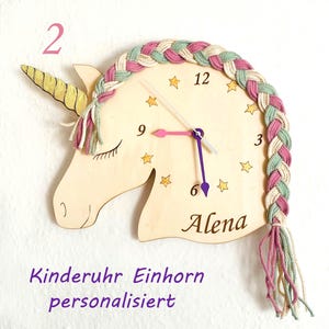 Könnte beinhalten: Eine hölzerne Einhorn-Wanduhr mit geflochtener Mähne in Rosa, Grün und Creme. Das Zifferblatt hat Sterne, schwarze Ziffern und den Namen "Alena". Das Einhornhorn ist gelb und die Zeiger sind rosa und lila.
