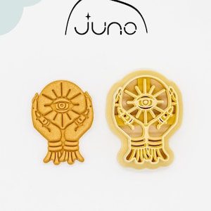 Puede incluir: Un cortador de galletas impreso en 3D en forma de dos manos sosteniendo una esfera con un ojo dentro. El cortador está hecho de plástico vegetal PLA y es no tóxico. El cortador es de color dorado.