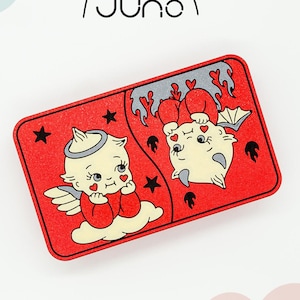 May include: A red rectangular card with a split image of an angel and a devil. The angel is on the left side of the card and is wearing a white halo and a red dress. The devil is on the right side of the card and is wearing a red dress and has horns and a tail. Both figures have hearts for eyes.