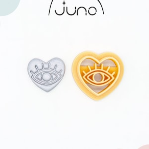 May include: Two 3D printed heart-shaped cookie cutters with an eye design. The cutter on the left is silver and the cutter on the right is light orange.