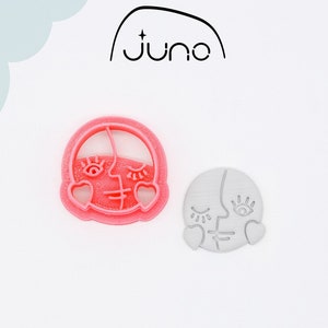 May include: Two 3D printed cookie cutters in the shape of a face with closed eyes and hearts. The cutter on the left is pink and the cutter on the right is gray. The cutters are made of PLA plant-based plastic, non-toxic.