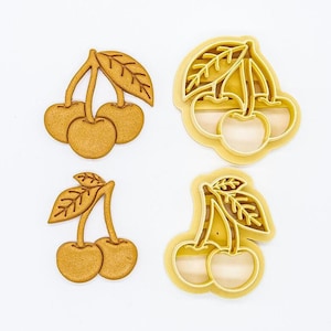 Puede incluir: Cuatro cortadores de galletas con forma de cerezas con hojas. Los cortadores están hechos de metal dorado.