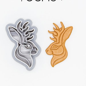 Puede incluir: Dos cortadores de galletas impresos en 3D, uno en gris y otro en marrón, con forma de cabeza de ciervo con cuernos. Los cortadores están hechos de plástico vegetal PLA y son no tóxicos.