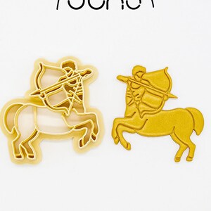 May include: Two gold-coloured Sagittarius zodiac sign cookie cutters. One is a 3D printed cutter, and the other is a solid gold-coloured Sagittarius. The cutters are made of PLA plant-based plastic, non-toxic. The logo "juno" is at the top.