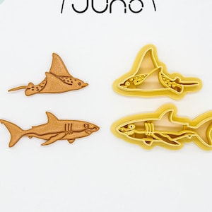 May include: Four shark and ray-shaped cookie cutters. Two are solid brown, and two are yellow outlines. The logo "juno" is in black script above the cutters. The cutters are on a white background.