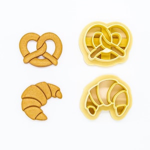 Puede incluir: Cuatro cortadores de galletas, dos con forma de pretzel y dos con forma de croissant. Los cortadores son amarillos y están hechos de plástico.
