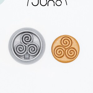 May include: A silver 3D printed cookie cutter in the shape of a triple spiral design. The cutter is shown next to a golden brown cookie with the same design.