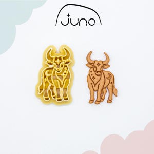 May include: Two Taurus-themed cookie cutters. One is a yellow plastic cutter, and the other is a brown cookie. The cookie cutters are in the shape of a bull with horns. The word "juno" is above the cookie cutters.