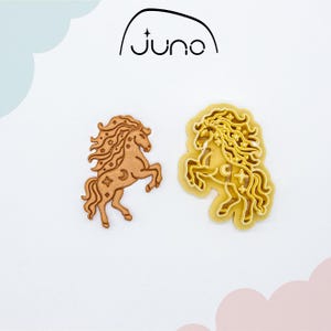 Könnte beinhalten: Zwei pferdeförmige Ausstecher. Einer ist hellbraun, der andere gelb. Beide haben ein himmlisches Design mit Sternen und Monden. Das Wort "juno" steht in einer schwarzen, stilisierten Schriftart über den Ausstechern.