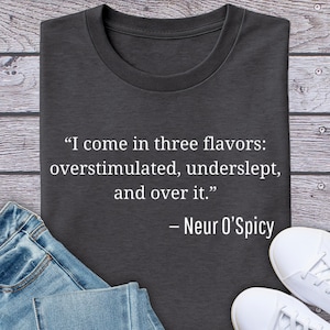 May include: Dark gray t-shirt with the quote "I come in three flavors: overstimulated, underslept, and over it." by Neur O'Spicy in white text. The shirt is folded on a wooden surface, with blue jeans and white sneakers visible.