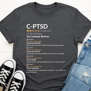 Può includere: T-shirt grigio scuro con la scritta "C-PTSD" e recensioni dei clienti. La maglietta ha un girocollo e maniche corte. Le scarpe da ginnastica bianche e nere sono visibili in basso a sinistra e un paio di jeans blu a destra.