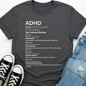 Puede incluir: Camiseta gris oscuro con la palabra "ADHD" y reseñas de clientes impresas. Las reseñas incluyen comentarios sobre "Never Bored", "Never Finishes" y "Random Wisdom". La camiseta se combina con vaqueros azules y zapatillas negras.