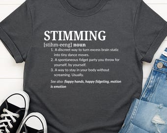 May include: Dark gray t-shirt with the word "STIMMING" in large white letters. The shirt includes a definition of the word, explaining it as a way to manage excess brain static. The shirt is paired with jeans and black and white sneakers.