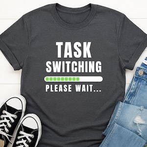 Può includere: T-shirt grigio scuro con la scritta bianca "TASK SWITCHING PLEASE WAIT...". Sotto il testo è presente una barra di avanzamento verde. Sono visibili scarpe da ginnastica bianche e nere e jeans blu.