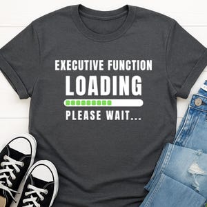 Puede incluir: Camiseta gris oscuro con el texto blanco "EXECUTIVE FUNCTION LOADING PLEASE WAIT...". La palabra "LOADING" tiene una barra de progreso verde. Zapatillas deportivas blancas y negras y vaqueros azules son visibles.