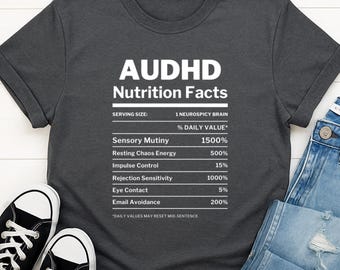 May include: Dark gray t-shirt with the text "AUDHD Nutrition Facts." The graphic lists humorous "daily values" for traits like "Sensory Mutiny" and "Email Avoidance." The shirt is paired with jeans and black sneakers.
