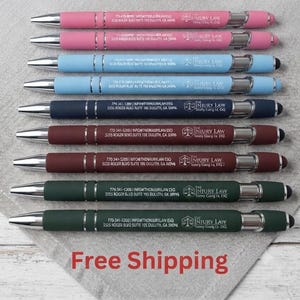May include: A set of nine retractable ballpoint pens in pink, light blue, navy blue, maroon, and dark green. Each pen features a silver clip, a stylus tip, and white printed text reading The Injury Law, contact details, and a scale of justice icon.