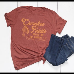 May include: A rust-colored t-shirt with the text "Cherokee Fiddle played for the whiskey" in orange. The text is in a vintage font and includes a stylized Native American headdress.