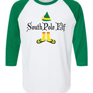 May include: White and green raglan style shirt with the text "South Pole Elf". The shirt features a green elf hat, yellow socks, and green and yellow sandals. The sleeves are green.