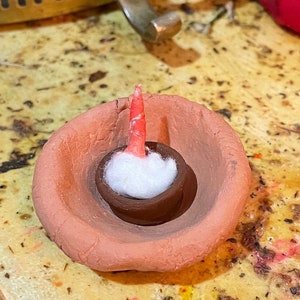May include: A small terracotta pot with a white cotton wick inside. The pot is sitting on a wooden surface.