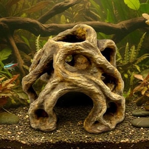 May include: A decorative aquarium ornament shaped like a weathered tree trunk with multiple openings. The brown and tan resin structure provides a naturalistic hiding place for fish. The background features aquatic plants and gravel.