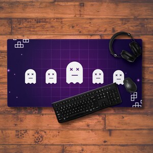 May include: A purple and black gaming mousepad with a grid pattern and five white ghost icons. The ghosts have a simple design with two eyes and a mouth. The mousepad also features a black keyboard and a black gaming headset.